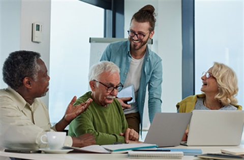 3 adults sitting around a desk, with one man standing behind them chatting