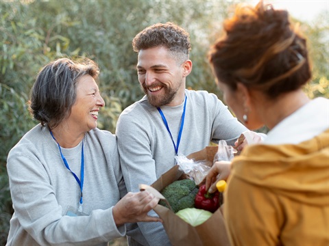 two people with a paper bag of produce smiling each other as they deliver it to another person