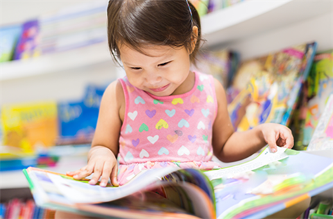 A young girl engrossed in a book, surrounded a shelf of picture books